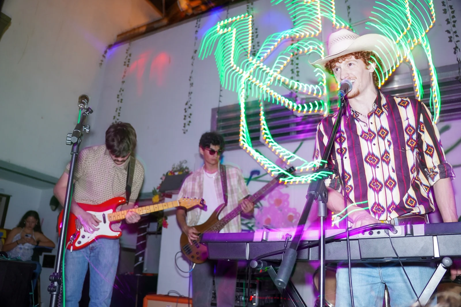 Miles smiling at the microphone behind a keyboard, surrounded by colored light trails and the band